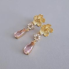 two gold earrings with pink stones and flowers on top of each earring, sitting on a white surface