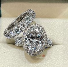 two diamond engagement rings sitting on top of a white cloth covered box with the ring in it's center surrounded by smaller diamonds