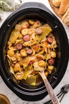 a crock pot filled with pasta and sausage next to bread on a marble table