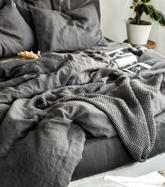 an unmade bed with dark gray linens and a potted plant in the corner