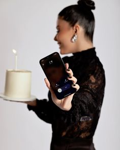 a woman holding a cell phone in front of a cake on a platter with a candle