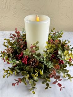 a lit candle surrounded by greenery and pine cones on a white tablecloth with a wall in the background