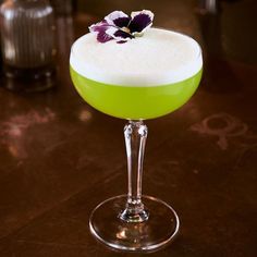 a green cocktail sitting on top of a table next to a glass with a flower in it