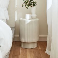 a white vase with flowers sitting on top of it next to a bed and window