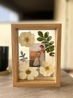 a wooden frame with white flowers and leaves on the table next to a black cup