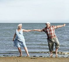 an older man and woman are holding hands on the beach by the water's edge