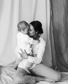 a woman holding a baby in her arms while sitting on top of a bed next to a curtain