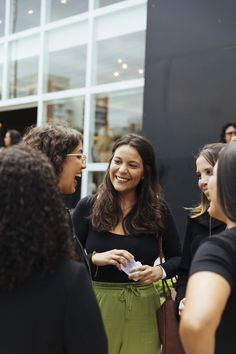 two women talking to each other in front of a building with many people standing around