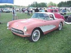 an old pink and white car parked in the grass at a car show with people standing around