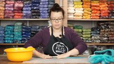 a woman in an apron is working at a table with bowls and yarns behind her