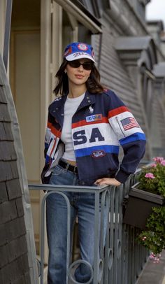 a woman standing next to a fence wearing a jacket and hat with an american flag on it