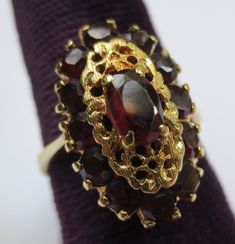 This ring looks so regal! The deep red garnets against the rich colored 18k gold compliment each other beautifully. Dating from the early 1900's-40's and hallmarked in the interior 750 and 176AAL. Has been tested to ensure the golds purity. Rings size is 6 1/2 and is ready to be worn and admired! The halo setting measures 11/16 by 7/16 of an inch. Stones are all held securely in place. Center garnet measures 6.12mm by 4.25mm. Some stones have light surface wear as to be expected with a ring of s Elegant Garnet Ruby Ring Stamped 14k, Formal Garnet Burgundy Ring, Formal Burgundy Garnet Ring, Formal Garnet Gemstone Cluster Ring, Victorian Gold Cluster Ring With Gemstone, Heirloom Garnet Cluster Ring For Anniversary, Antique Yellow Gold Ruby Ring With Polished Finish, Vintage Red Garnet Cluster Ring, Classic Burgundy Ruby Ring For Formal Occasions