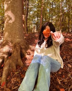 a woman sitting on the ground next to a tree holding an orange object in front of her face