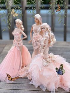 three barbie dolls in pink dresses on a wooden platform with butterflies around them and trees behind them