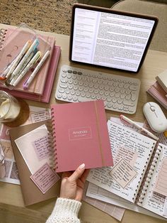 a person holding a pink notebook next to a keyboard and laptop computer on a desk