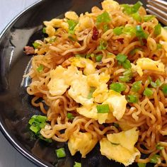 a black plate topped with noodles and vegetables on top of a white table next to a fork