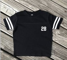 This adorable T-shirt is the perfect outfit to accompany dad while watching his favorite sport. Unique personalized children's gift. Please provide child's name or last name & desired number on the note to seller portion at checkout. Available colors: Black Smoke Gray Green Pink Royal Blue Red White Please note: due to variations in computer monitors & phone screens there might be a slight difference in color between the product shown and the actual item** ⚽️⚽️⚽️⚽️⚽️⚽️⚽️⚽️⚽️⚽️⚽️⚽️⚽️⚽️⚽️⚽ Black Varsity T-shirt With Text Print, Sporty White T-shirt With Number Print, Black Varsity T-shirt For Game Day, Black T-shirt With Letter Print For Game Day, Black Number Print Top For Game Day, Short Sleeve T-shirt For Baseball Season With School Spirit, Black Tops With Name Print For Baseball Season, Black T-shirt With Football Season Text Print, Black T-shirt With Text Print For Football Season