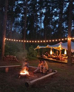 people sitting around a campfire in the woods at night with lights strung over them