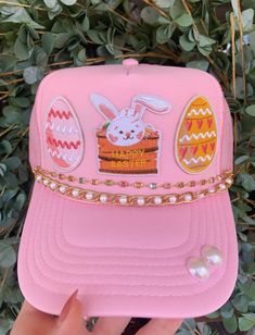 a pink hat with an embroidered bunny and eggs on it, sitting in front of some bushes