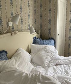 an unmade bed with white sheets and blue pillows in a room that has floral wallpaper on the walls