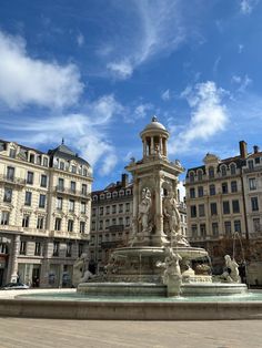 a fountain in the middle of a city square with buildings around it and blue skies above