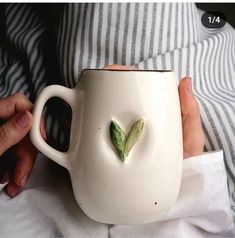 a person holding a white coffee mug with a green leaf in the shape of a heart