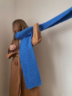 a woman wearing a blue knitted scarf with her arms wrapped around her head, standing in front of a white wall