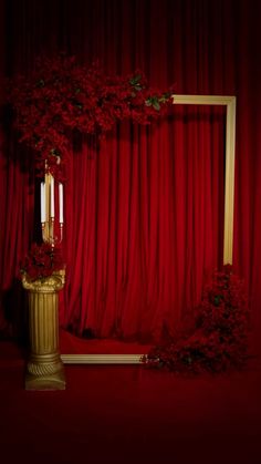 a gold vase with red flowers and two candles in front of a red velvet curtain