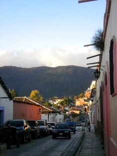 cars parked on the side of a road next to buildings and mountains in the distance