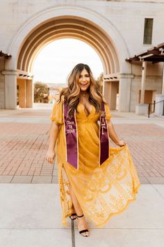 a woman in a yellow dress and purple sash standing on the sidewalk with an archway behind her