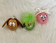 three ornaments with faces painted on them sitting on a white fur covered surface and one has a green fuzzy ball in the shape of a dog's head