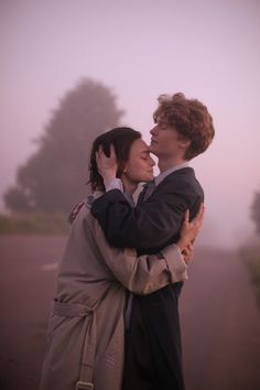 two people standing next to each other in front of a foggy road and trees