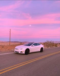 a white car driving down the road in front of a pink sky and some telephone poles