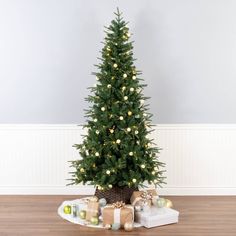 a small christmas tree with lights and presents under it on a wooden floor in front of a white wall