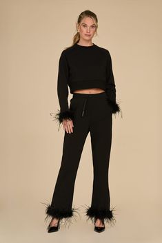 A woman gracefully wears the Rada Feather Trim Pant - Black, featuring stylish ostrich feather trim on the sleeves and pant legs. Posed against a simple beige background, she pairs her ensemble with sleek black heels and sports a chic half-up hairstyle, effortlessly radiating charm. Chic Evening Bottoms With Feathers, Chic Feathered Bottoms For Evening, Chic Feather Trim Bottoms For Night Out, Chic Feather Trim Bottoms, Chic Pants With Feather Trim, Feather Trim Party Bottoms For Fall, Ostrich Feather Trim, Feather Trim, Ostrich Feather