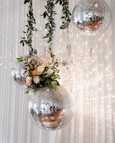 three disco ball vases with flowers and greenery hanging from the ceiling in front of a white curtain