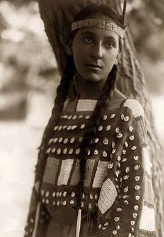 an old black and white photo of a woman in native american clothing standing next to a tree