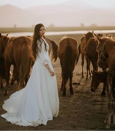 a woman in a white dress standing next to some brown horses on a dirt field