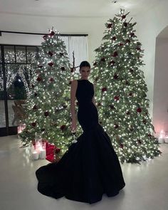 a woman standing in front of a christmas tree wearing a black evening gown and matching heels