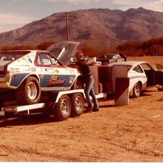 two men are working on the back of a truck in the middle of dirt with mountains in the background