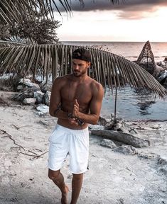 a shirtless man walking on the beach with his hands clasped to his chest while wearing white shorts