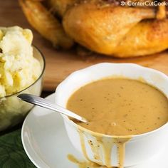 a bowl of soup next to a plate of food on a wooden table with bread and croissants in the background