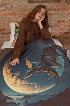 a woman sitting on top of a bed covered in a dragon blanket with the moon behind her