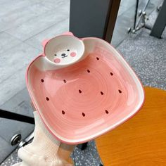 a person holding a pink plate with a teddy bear on it's side and a white bowl in the shape of a watermelon