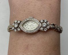 Absolutely stunning women's wrist watch is made of 14 karat white gold and has 67 small round diamonds. The watch is a Hamilton Registered wrist watch and comes in its original red felt box. The back has a locking clasp and a safety chain. There are 13 diamonds on one side of the band and 16 on the other side of the band. The face of the watch has 20 diamonds framing it, and there are 2 flowers made of 7 diamonds each on the sides of the face, and 2 small diamonds in between. The 14 diamonds in the flowers are slightly larger than all the rest.  Condition: Excellent! Stamped/Hallmarked: 14K Gold ©️ Hamilton (on back of watch case)   -14K- (on clasp) Weight: 22.5 grams  Measurements: 7 1/4 inches around inside (when closed) *Case: 7/8 inch long 5/8 inch wide *Face: 5/8 inch long 3/8 inch wi Diamond Anniversary Watch With Jubilee Bracelet, Diamond Watch With Jubilee Bracelet For Anniversary, Diamond Watch With 17 Jewels As Gift, White Gold Diamond Watch With Jubilee Bracelet For Anniversary, Wedding Diamond Watch With Jubilee Bracelet, Single Cut Diamond Jewelry And Watches, Round Watches With Single Cut Diamonds For Anniversary, Anniversary Watches With Single Cut Diamonds, Anniversary Diamond Watch With Single Cut Diamonds