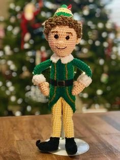 a crocheted elf is standing in front of a christmas tree with his hands on his hips