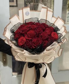 a bouquet of red roses wrapped in white paper and tied with black ribbon, held by someone's hand