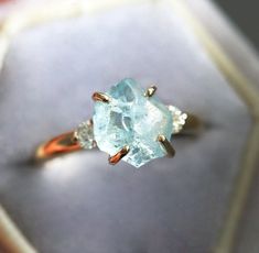 Raw gemstone ring with accented diamonds. Please select the main stone from the drop down menu.  𝗗𝗘𝗧𝗔𝗜𝗟𝗦 𝗠𝗔𝗜𝗡 𝗦𝗧𝗢𝗡𝗘 𝗚𝗲𝗺𝘀𝘁𝗼𝗻𝗲: Aquamarine 𝗖𝗼𝗹𝗼𝗿: Blue 𝗦𝗵𝗮𝗽𝗲: Other 𝗖𝘂𝘁: Raw 𝗠𝗲𝗮𝘀𝘂𝗿𝗲𝗺𝗲𝗻𝘁: 9x8 mm 𝗦𝗜𝗗𝗘 𝗦𝗧𝗢𝗡𝗘𝗦 𝗗𝗶𝗮𝗺𝗼𝗻𝗱, round, brilliant cut, 3.00 mm (approx. 0.22 ct) (2) 𝗗𝗶𝗮𝗺𝗼𝗻𝗱 𝗾𝘂𝗮𝗹𝗶𝘁𝘆: Clarity grade VS, color grade F-G, conflict-free 𝗕𝗔𝗡𝗗 𝗦𝘁𝘆𝗹𝗲: 3-stone 𝗠𝗮𝘁𝗲𝗿𝗶𝗮𝗹: 14k gold (yellow, white, rose), 18k gold (ye Engagement Ring With Side Diamonds, Raw Stone Engagement Rings, Three Stone Diamond Rings Engagement, Gemstone Engagement Ring, Raw Gemstone Ring, Aquamarine Engagement Ring, Topaz Color, Three Stone Diamond, Gemstone Engagement