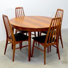 a wooden table and four chairs with black cushions on the bottom, against a white wall