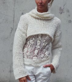 a woman standing in front of a concrete wall wearing a white sweater and white pants